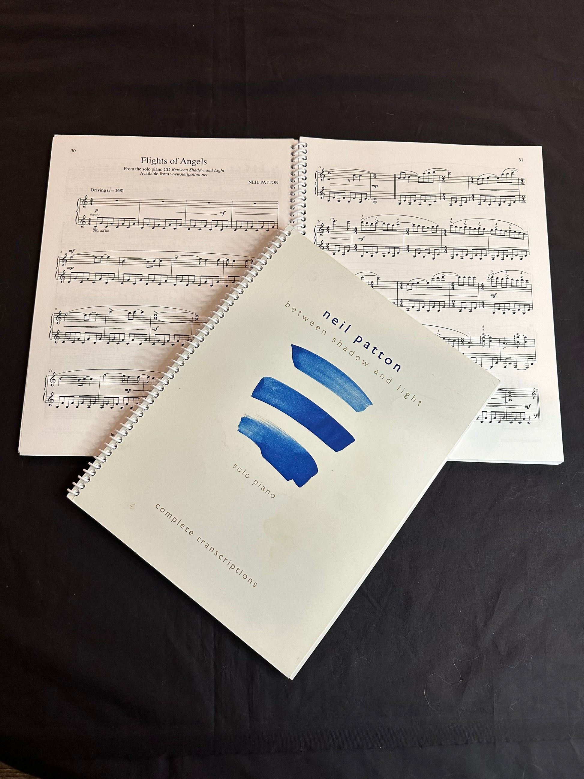 A closed copy of the Between Shadow and Light Songbook lying on another open copy, displaying a sample of the printed music inside.