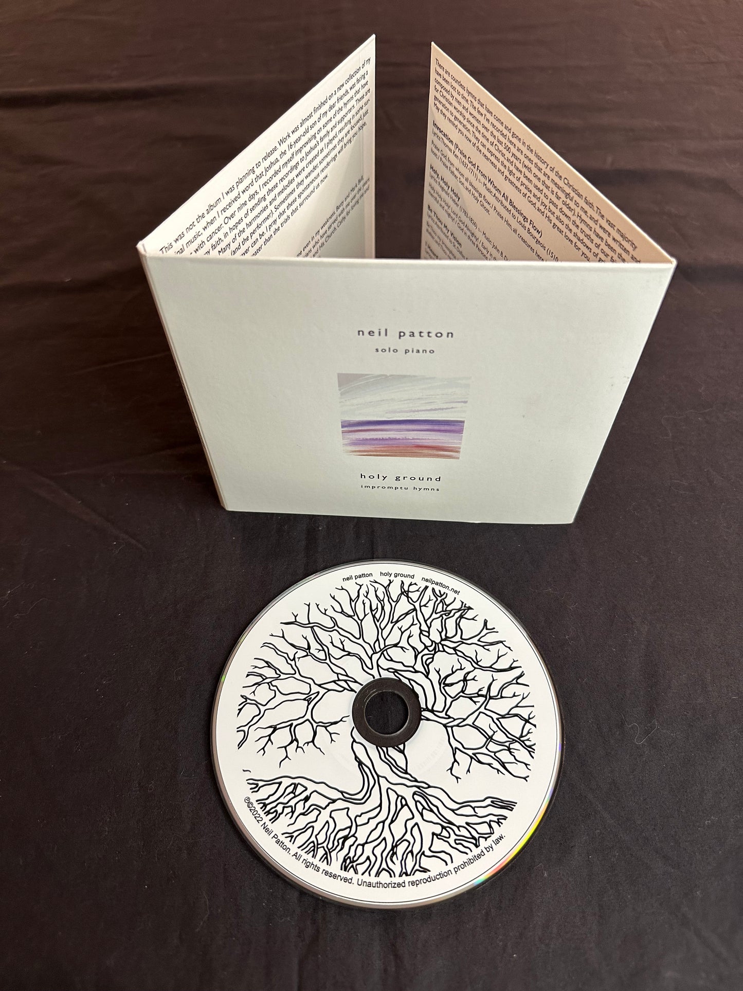 The Holy Ground Compact Disc lies in the foreground with an image of a tree and roots on it, next to the cardboard package for the album, showing the front cover and some of the inner liner notes.