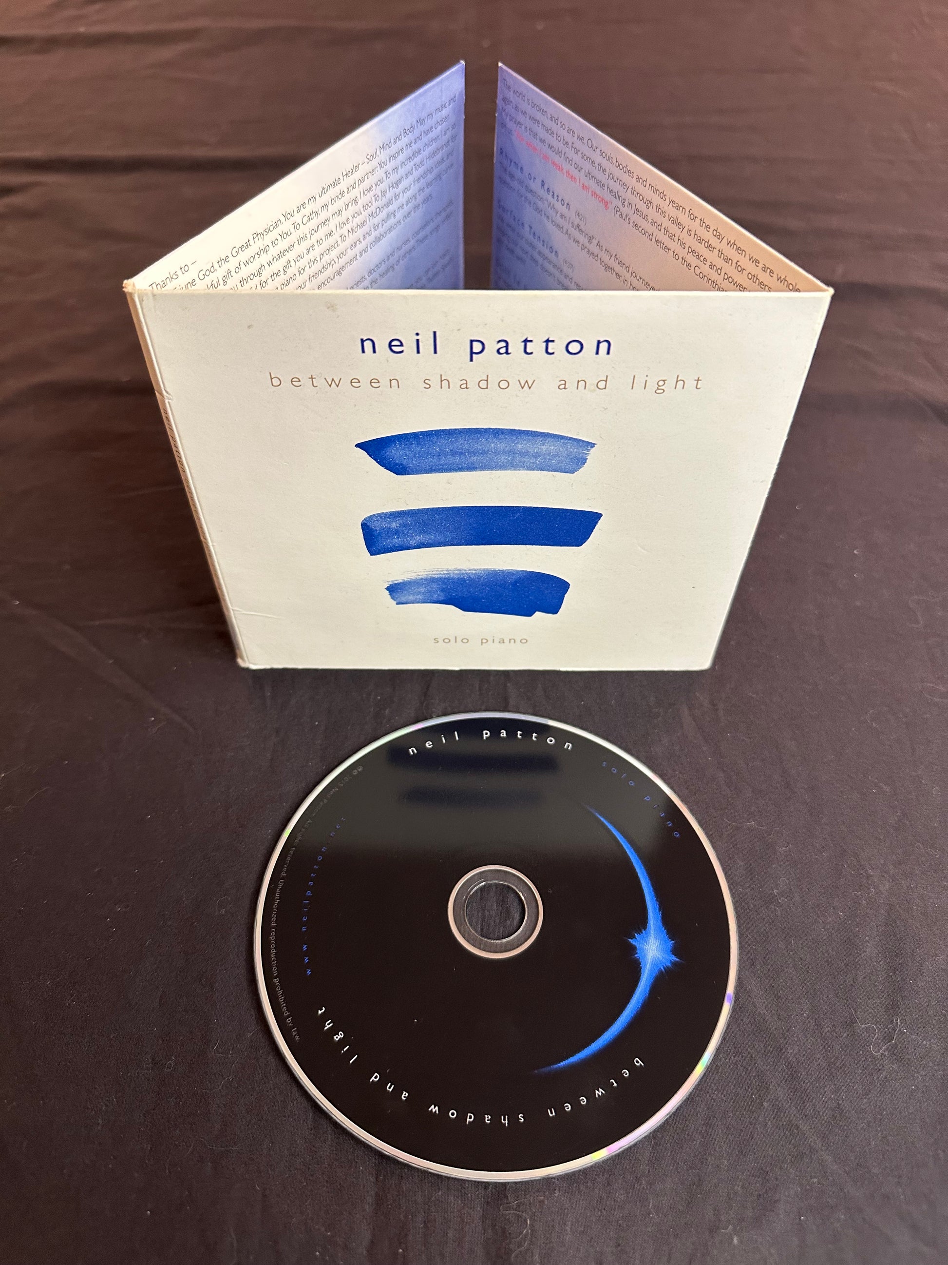 The Between Shadow and Light comp[act Disc lying in front of its cardboard case showing the front cover and some fo the text from the inside liner notes.